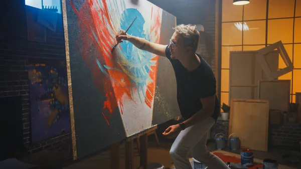 Artista talentoso que trabaja en la pintura abstracta, utiliza el pincel de pintura para crear una imagen moderna audazmente emocional. Estudio creativo oscuro grandes soportes de lona en caballete iluminado . —  Fotos de Stock