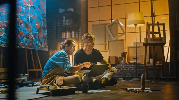 Un par de artistas sentados en su estudio usan un ordenador portátil, trabajan en un proyecto, hablan y sonríen. Painter and Her Partner Doing Research (en inglés). Taller auténtico con pinturas al óleo y herramientas — Foto de Stock