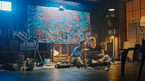Un par de artistas sentados en su estudio usan un ordenador portátil, trabajan en un proyecto, hablan y sonríen. Painter and Her Partner Doing Research (en inglés). Taller auténtico con pinturas al óleo y herramientas — Foto de Stock