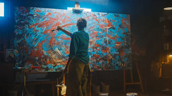 Utalentowana kobieta artysta pracuje na abstrakcyjne malowanie olejowe, za pomocą farby Pędzel tworzy Modern arcydzieło. Ciemne i brudne Studio kreatywne, gdzie duże płótno stoi na Easel podświetlane. — Zdjęcie stockowe