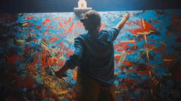 Talentosa artista femenina trabaja en la pintura al óleo abstracta, utilizando el pincel de pintura que crea obra maestra moderna. Estudio creativo oscuro y desordenado donde el lienzo grande se destaca en el caballete iluminado . — Foto de Stock