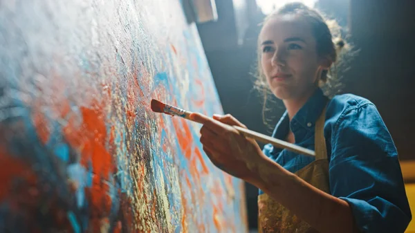 Artista femenina trabaja en la pintura al óleo abstracta, Pincel en movimiento Enérgicamente crea obra maestra moderna. Dark Creative Studio donde Large Canvas se destaca en el caballete iluminado. Primer plano de ángulo bajo — Foto de Stock