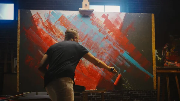 Талантливый мужчина-художник, работающий над абстрактной живописью, использует промышленный ролик для создания смелой эмоциональной современной картины. Тёмная творческая студия Large Canon стоит на иллюминированной простыне — стоковое фото