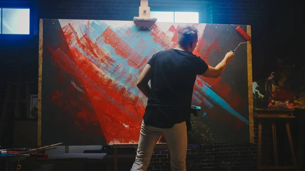 Talentoso artista masculino que trabaja en una pintura abstracta, utiliza un rodillo industrial para crear una imagen moderna audazmente emocional. Estudio creativo oscuro grandes soportes de lona en caballete iluminado — Foto de Stock