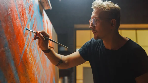 Artista talentoso que trabaja en la pintura abstracta, se sumerge en la pintura y comienza a dibujar arte moderno audazmente emocional. Estudio creativo oscuro grandes soportes de lona en caballete iluminado — Foto de Stock