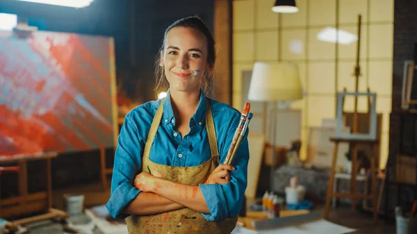 Jovem artista feminina profissional sujo com pintura, vestindo avental, braços cruzados enquanto segura escovas, olha para a câmera com um sorriso. Authentic Creative Studio com tela grande. Retrato facial — Fotografia de Stock