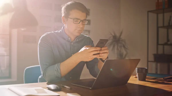 Professionelle kreative Mann sitzt an seinem Schreibtisch im Homeoffice-Studio und arbeitet an einem Laptop. Mann nutzt Smartphone, um soziale Medien zu checken, Internet zu durchsuchen. — Stockfoto