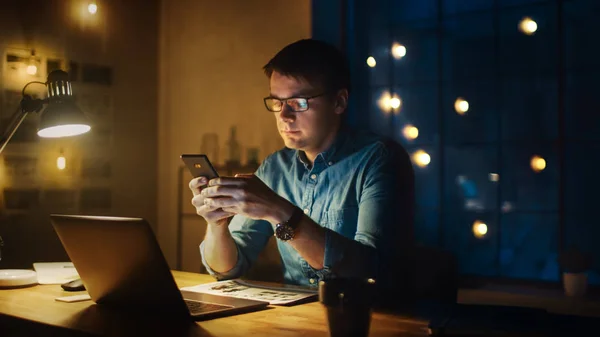 Profesionální muž sedí za stolem v Office Studiu a večer pracuje na laptopu. Muž používající smartphone s daty, analýza statistiky. — Stock fotografie