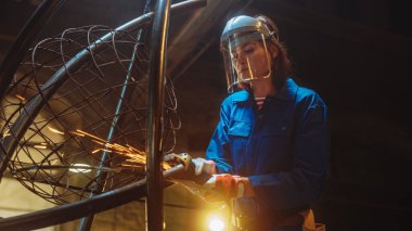 Mavi Tulumlu Genç Çağdaş Kadın Sanatçı, Stüdyo atölyesinde Bir Angle Grinder ile Metal Metro Heykeli Öğütüyor. Güçlendirici Kadın Metalden Modern Vahşi Sanatsal Çalışmalar Yapıyor.