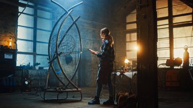 Güzel Kadın Sanatçı, Brutal Metal Heykeli Stüdyodaki Tablet Skeç 'le karşılaştırıyor. Erkek Fatma Kız Kareli Gömlek ve Önlük Giyiyor. Çağdaş Üretici Soyut Çelik Sanatı Oluşturuyor.