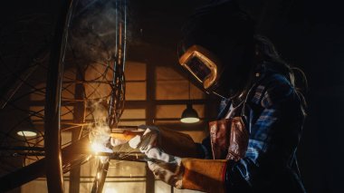 Güzel Kadın Sanatçı Karanlık Stüdyoda Zalim Metal Heykel Kaynağı. Erkek Fatma Kız, üzerinde kıvılcımlar uçuşan metal tüp parlatıyor. Çağdaş Üretici Soyut Çelik Sanatı Oluşturuyor.