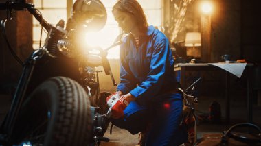 Genç Güzel Kadın Tamirci, Özel Bobber Motosikleti üzerinde çalışıyor. Mavi Tulum Giyen Yetenekli Kız. Fındık cıvatalarını sıkmak için bir İngiliz anahtarı kullanıyor. Yaratıcı Otantik Atölye Garajı.