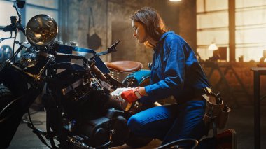 Young Beautiful Female Mechanic Comes Working on a Custom Motorcycle in Garage. Talented Girl Wearing a Blue Jumpsuit. She Uses a Ratchet to Tighten Nut Bolts. Creative Authentic Workshop. clipart