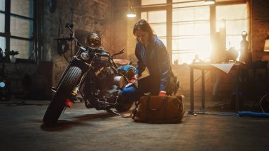 Young Beautiful Female Mechanic Comes Working on a Custom Motorcycle in Garage. Talented Girl Wearing a Blue Jumpsuit. She Looking for an Instrument in a Bag. Creative Authentic Workshop. clipart