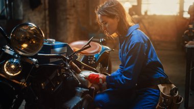 Young Beautiful Female Mechanic is Working on a Custom Bobber Motorcycle. Talented Girl Wearing a Blue Jumpsuit. She Uses a Ratchet Spanner to Tighten Nut Bolts. Creative Authentic Workshop Garage. clipart