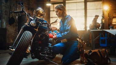 Genç Güzel Kadın Tamirci, Özel Bobber Motosikleti üzerinde çalışıyor. Mavi Tulum Giyen Yetenekli Kız. Fındık cıvatalarını sıkmak için Ratchet anahtarı kullanıyor. Yaratıcı Otantik Atölye Garajı.