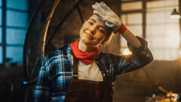Young Beautiful Empowering Woman Rubs Her Forehead and Gently Smiles at the Camera. Authentic Fabricator Wearing Work Clothes in a Metal Workshop.
