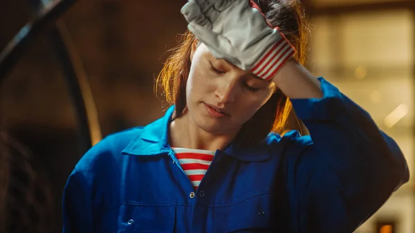 Young Beautiful Empowering Woman Rubs Her Forehead. Authentic Fabricator Wearing Work Clothes in a Metal Workshop. Sparks Flying on the Background.