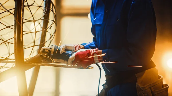 Close-up van jonge vrouwelijke Fabricator in blauw jumpsuit. Ze slijpt een metalen buizensculptuur met een hoekslijper in een atelier. Empowering Woman maakt moderne abstracte metalen kunstwerken. — Stockfoto