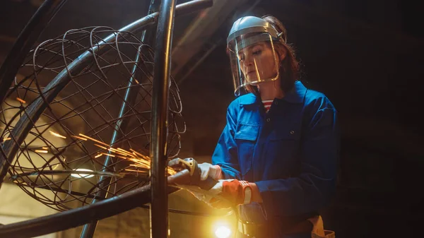 Junge zeitgenössische Künstlerin im blauen Overall schleift in einer Atelierwerkstatt eine Metallrohrskulptur mit einem Winkelschleifer. Empowering Woman macht moderne brutale abstrakte Kunstwerke aus Metall. — Stockfoto