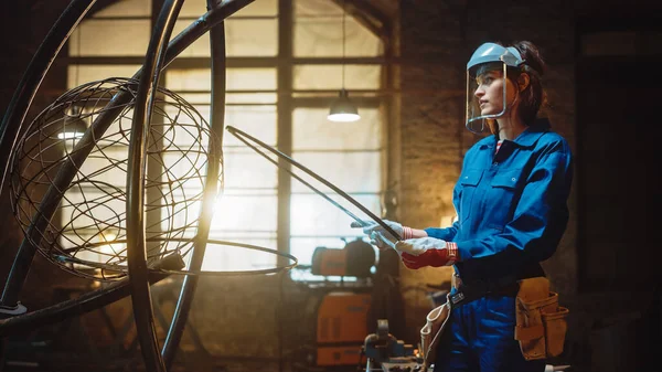 Young Contemporary Female Artist in Blue Jumpsuit and Safety Mask monteert een metalen ring op een buizensculptuur in een atelieratelier. Empowering vrouw maakt moderne brutale abstracte kunst uit staal. — Stockfoto