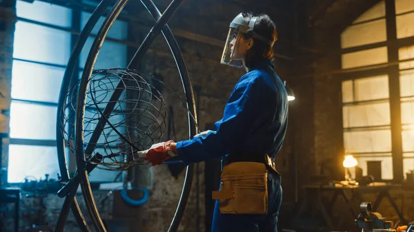 Молода сучасна жінка-виконавиця в Blue Jumpsuit and Safety Mask є Fitting a Metal Ring to a Tube Sculpture в студії Workshop. Підсилююча жінка витворює сучасні брутальні абстрактні витвори мистецтва. — стокове фото