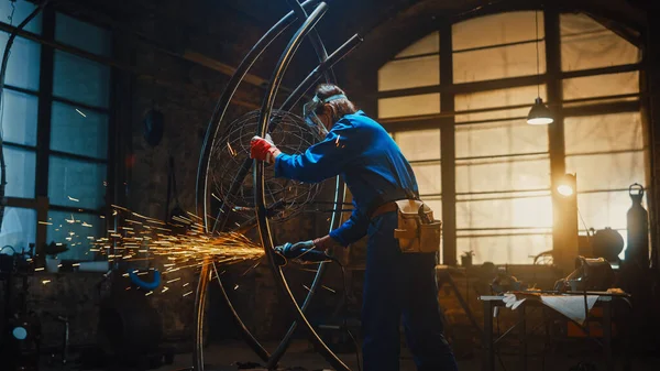 Joven artista femenina contemporánea en mono azul está moliendo una escultura de tubo de metal con una amoladora angular en un taller de estudio. Empoderar a la mujer hace obras de arte abstractas brutales modernas de metal . — Foto de Stock