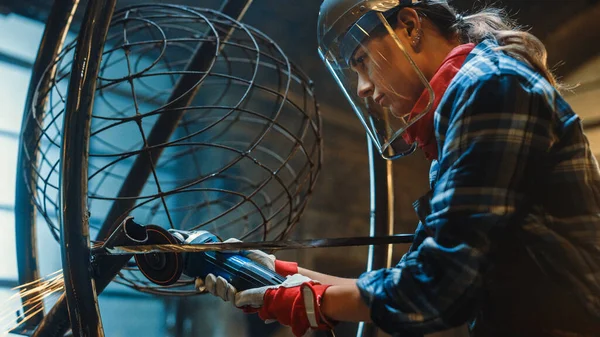 Primer plano de la joven fabricadora en máscara de seguridad. Ella está moliendo una escultura de tubo de metal con una amoladora angular en un taller de estudio. Empoderar a la mujer hace obras de arte de metal abstracto moderno . — Foto de Stock