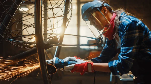 Primer plano de la joven fabricadora en máscara de seguridad. Ella está moliendo una escultura de tubo de metal con una amoladora angular en un taller de estudio. Empoderar a la mujer hace obras de arte de metal abstracto moderno . — Foto de Stock