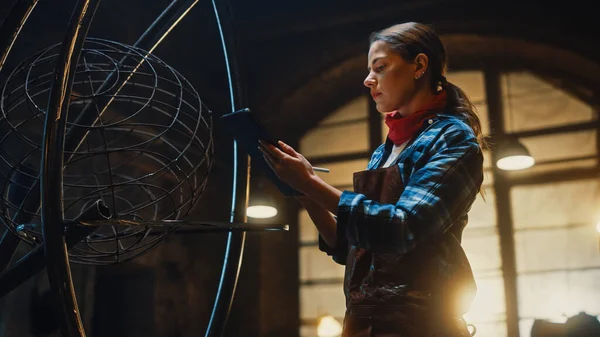 Hermosa artista femenina bocetos en una tableta al lado de la escultura de metal brutal en el estudio. Tomboy Girl lleva camisa a cuadros y delantal. Fabricador contemporáneo Creación de arte abstracto de acero . — Foto de Stock