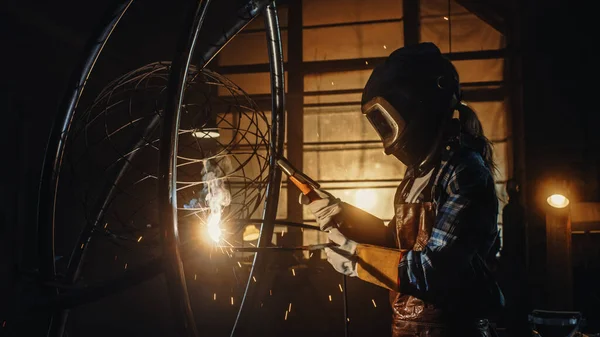 美丽的女艺术家正在黑暗工作室焊接一个残忍的金属雕塑。汤姆男孩女孩抛光金属管与火花飞出它。当代造型师创造抽象钢艺术. — 图库照片