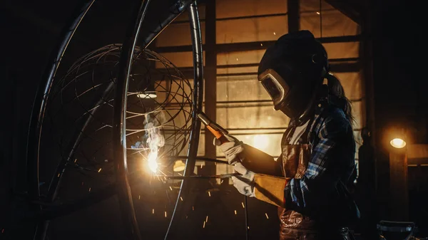 Piękna artystka spawa brutalną metalową rzeźbę w ciemnym studiu. Laska poleruje metalową rurkę z odlatującymi iskrami. Współczesny producent Tworzenie abstrakcyjnej sztuki stalowej. — Zdjęcie stockowe