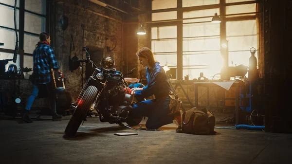 Jovem bela mecânica feminina está trabalhando em uma motocicleta Bobber personalizado. Garota talentosa vestindo um macacão azul. Ela usa uma chave-inglesa para apertar parafusos de porca. Creative Authentic Workshop Garagem . — Fotografia de Stock