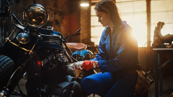 Joven hermosa mecánica femenina está fijando una motocicleta de bobina personalizada. Chica talentosa usando un mono azul. Usa una llave de trinquete. Taller creativo auténtico Garaje — Foto de Stock