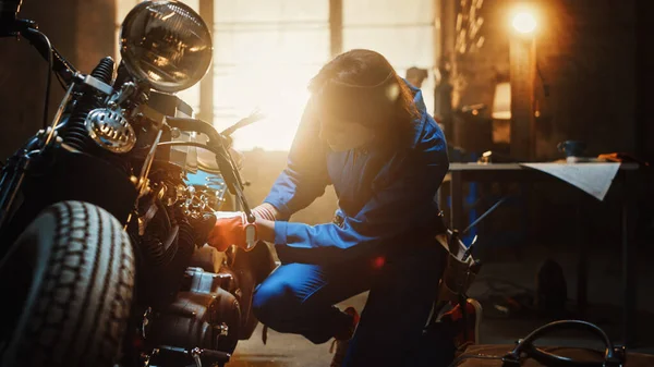 Ung vacker kvinnlig mekaniker arbetar på en anpassad Bobber motorcykel. En begåvad tjej i blå jumpram. Hon använder en Spanner för att dra åt mutterskruvarna. Kreativ autentisk verkstad Garage. — Stockfoto