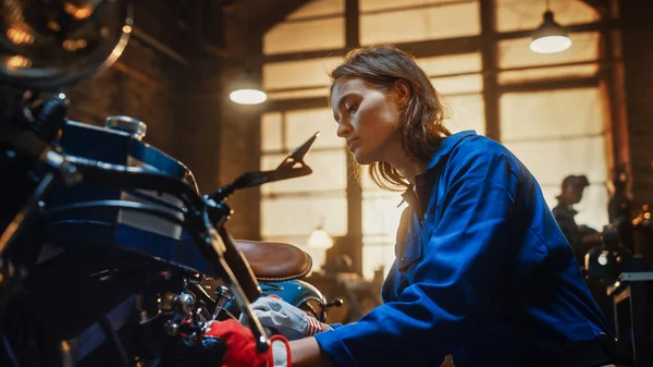 Joven hermosa mecánica femenina está trabajando en una motocicleta Bobber personalizado. Chica talentosa usando un mono azul. Usa una llave para apretar los tornillos de tuerca. Taller creativo auténtico Garaje . —  Fotos de Stock