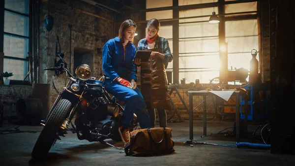 Két fiatal gyönyörű nő beszélget a munka végzett egyéni Bobber motorkerékpár. A tehetséges lányok táblagépet használnak. Ők jó szelleműek és boldogok. Creative Authentic Workshop Garázs. — Stock Fotó