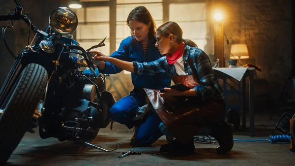 Duas jovens mulheres bonitas estão discutindo o trabalho feito em uma motocicleta Bobber personalizado. Garotas talentosas usam um computador tablet. Eles estão em bom espírito e feliz. Creative Authentic Workshop Garagem . — Fotografia de Stock
