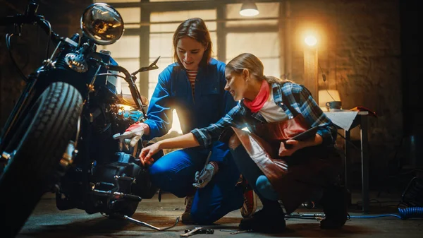 Dos jóvenes hermosas hembras están discutiendo el trabajo hecho en una motocicleta de bobina personalizada. Las chicas talentosas usan un Tablet Computer. Ellos están en buen espíritu y felices. Taller creativo auténtico Garaje . —  Fotos de Stock