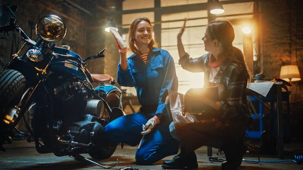 Két fiatal gyönyörű nő beszélget a munka végzett egyéni Bobber motorkerékpár. A tehetséges lányok táblagépet használnak. Boldogok és pacsiznak. Creative Authentic Workshop Garázs. — Stock Fotó