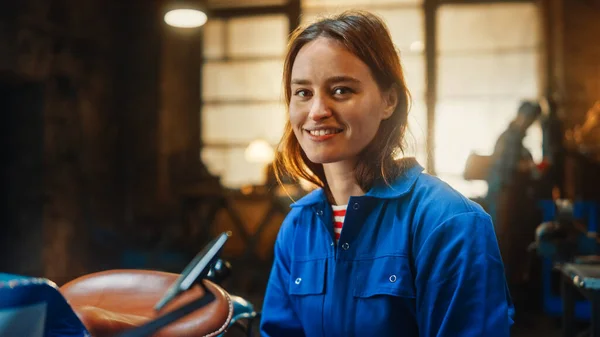 Jeune belle autonomisation femme sourit doucement à la caméra. Fabricant authentique portant des vêtements de travail dans un atelier de métal. — Photo