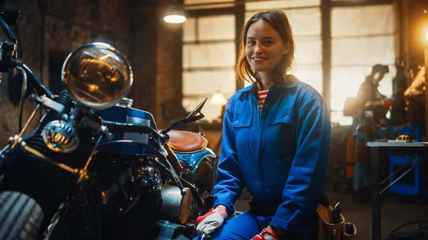 Joven hermosa mecánica femenina está trabajando en una motocicleta Bobber personalizado. Chica talentosa usando un mono azul. Ella sonríe a la cámara. Taller creativo auténtico Garaje . — Foto de Stock