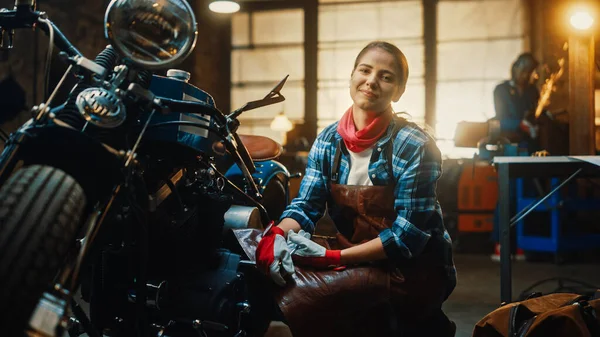 Młoda piękna kobieta mechanik pracuje na Custom Bobber Motocykl. Utalentowana dziewczyna w szachownicy i fartuchu. Uśmiecha się do kamery. Warsztaty Creative Authentic Garaż. — Zdjęcie stockowe
