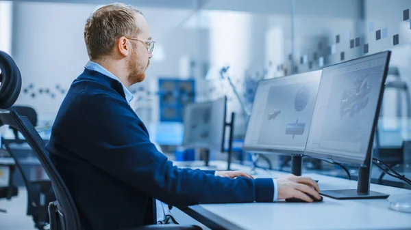 Ingeniero industrial Trabajando en una computadora personal con dos monitores mostrando software CAD con prototipo 3D de motor eléctrico híbrido y gráficos. Fábrica moderna con maquinaria de alta tecnología — Foto de Stock