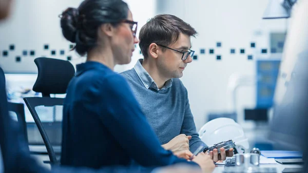 Modern Factory: El ingeniero industrial masculino explica a la supervisora del proyecto femenino las funciones de la parte de la máquina comparándola con una en la pantalla de la computadora. Oficina de fábrica industrial moderna de alta tecnología . — Foto de Stock