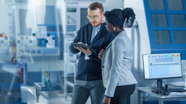 Kadın Endüstri Mühendisi Erkek Proje Yöneticisi, Tablet Bilgisayar kullanıyor ve Modern Fabrika Planlama 'yı tartışıyorlar. CNC Makineleri ve Robot Silahlanma Hattı ile Üretim Tesisi