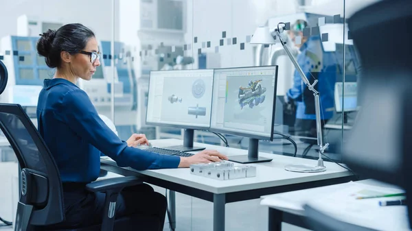 Female Industrial Engineer Solving Problems, Working on a Personal Computer, Two Monitor Screens Show CAD Software with 3D Prototype of Engine Being Tested. Working Modern Factory