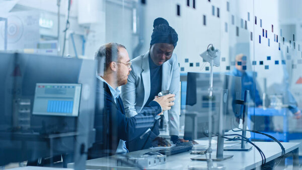 Современная фабрика: женщина-руководитель проекта разговаривает с мужским промышленным инженером, который работает на компьютере. Они улучшают механику и конструкцию машин. Рабочий высокотехнологичный промышленный комплекс
