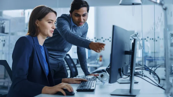 Modern Factory Office: Male Project Supervisor habla con una ingeniera industrial que trabaja en la computadora. Trabajo en equipo profesional, jóvenes especialistas solucionando problemas e impulsando el progreso tecnológico — Foto de Stock