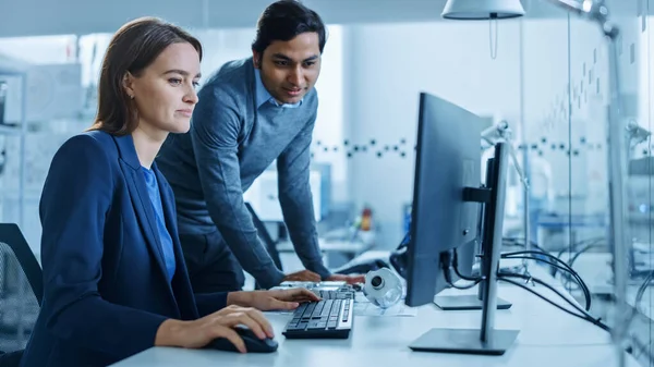 Modernes Fabrikbüro: Männlicher Projektleiter spricht mit einer Industrieingenieurin, die am Computer arbeitet. Professionelle Teamarbeit, junge Spezialisten, die Probleme lösen und den technologischen Fortschritt vorantreiben — Stockfoto